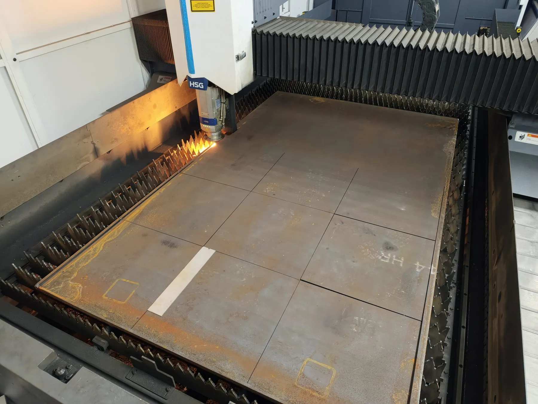 Table de la machine de découpe laser HSG vue de dessus : feuille de tôle avec contours de pièces découpés, étincelles près de la tête, soufflets de protection sur les côtés.