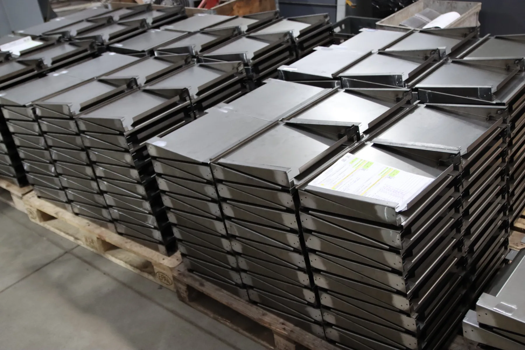 Piles de pièces en tôle pliées sur des palettes, avec la fiche de suivi de commande posée sur le dessus.