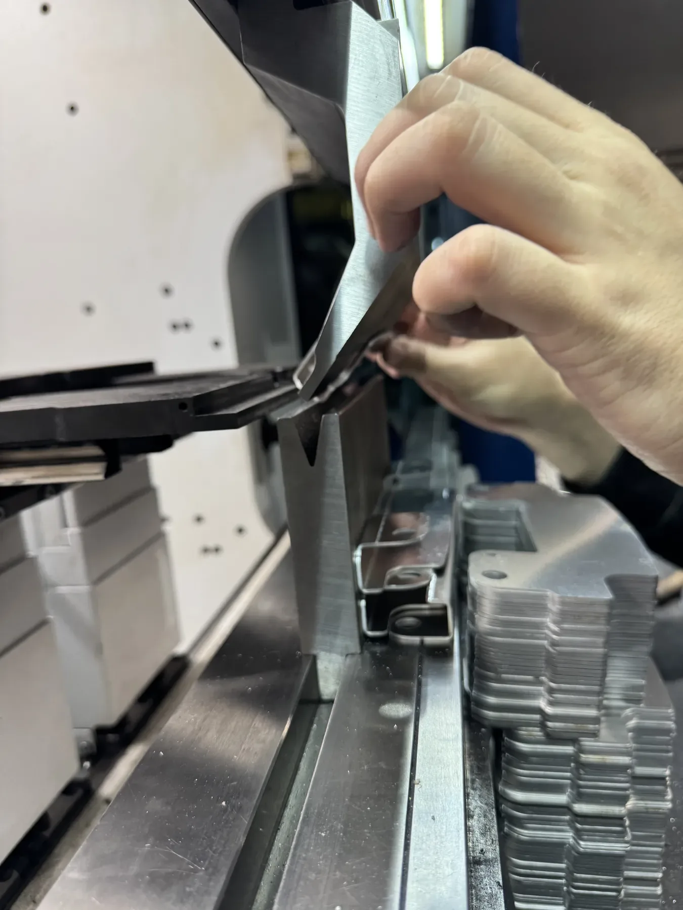 Main maintenant une tôle en cours de pliage dans l'outil de la presse plieuse : poinçon au-dessus de la matrice en V, pièce pliée à pattes et pile de flans.