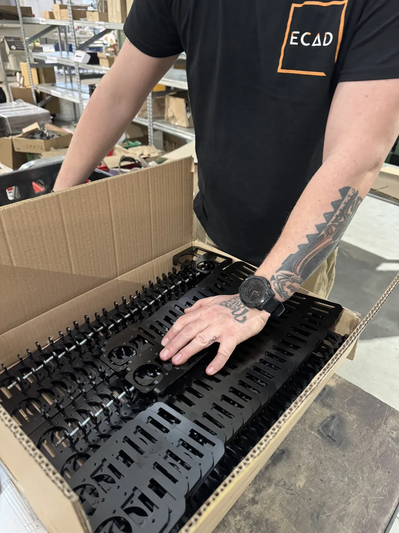 Employé en t-shirt ECAD rangeant des pièces noires en tôle dans un carton.