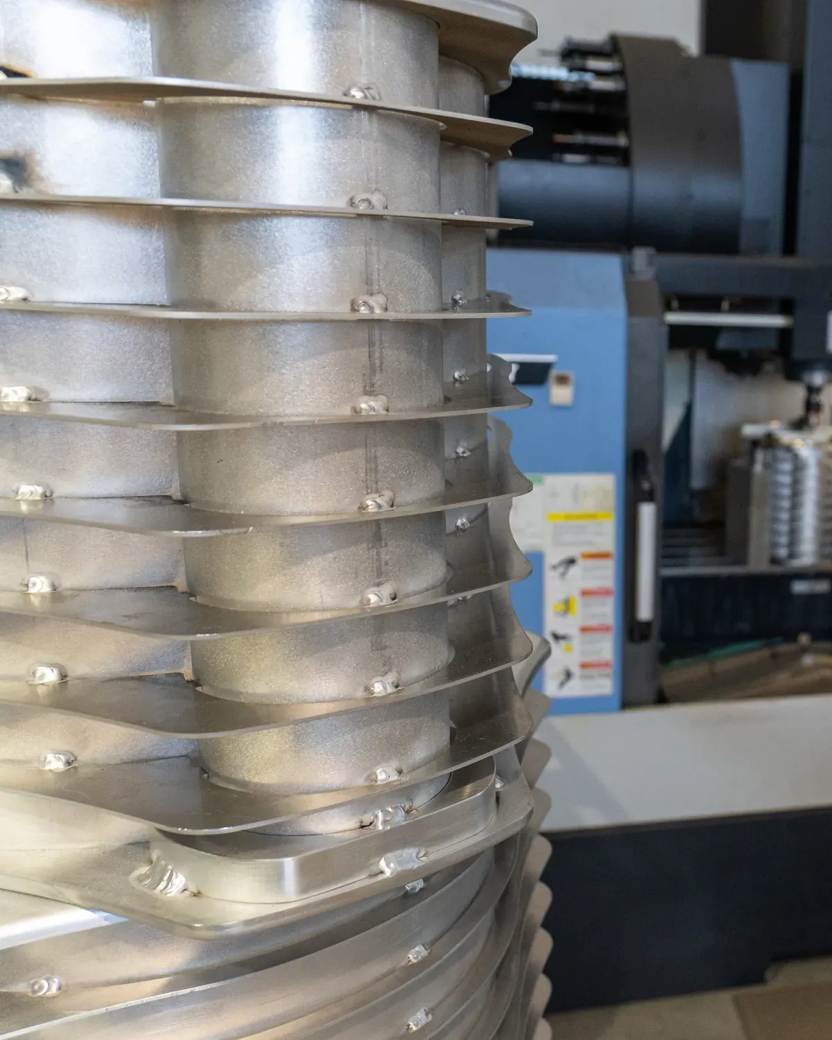 Pile de pièces en aluminium nervurées au premier plan, devant un centre d'usinage bleu avec sa commande numérique.